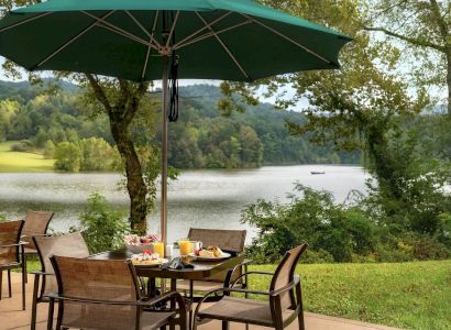 An outdoor dining setup under a green umbrella overlooks a scenic lake and wooded hills in the background. It ends the sentence.