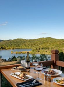 A table with food, wine, and cutlery is set on a deck overlooking a scenic lake and wooded hills under a clear blue sky.