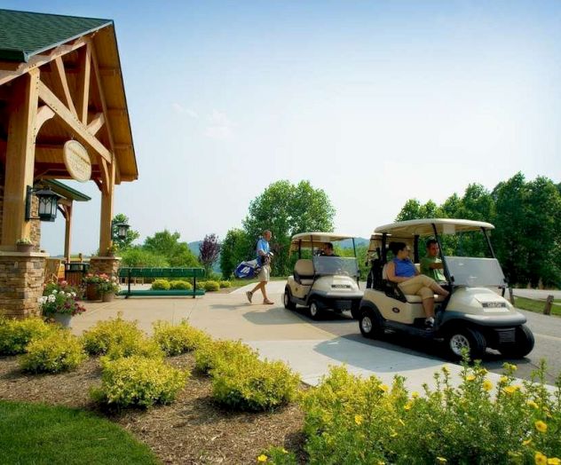 The image shows two people in golf carts near a building with wooden accents, while a third person walks towards them carrying golf clubs.