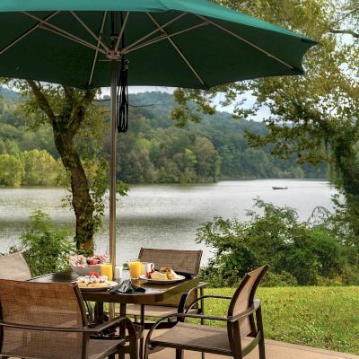 A table with a green umbrella, set for breakfast, overlooks a peaceful lake surrounded by trees and greenery in the background.