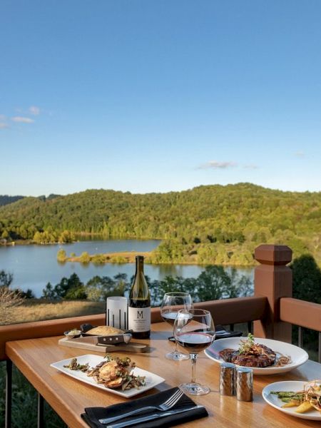 A dining table with plates of food, wine glasses, and a bottle, set on a balcony overlooking a scenic lake and forested hills during the daytime.