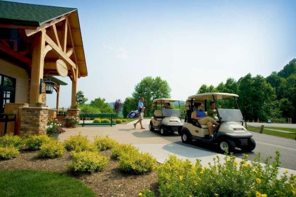 People are preparing for or returning from a golf outing near a clubhouse, with golf carts and sports equipment visible in a sunny setting.