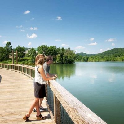 Two people are standing on a wooden boardwalk overlooking a calm lake surrounded by greenery and hills on a clear, sunny day.