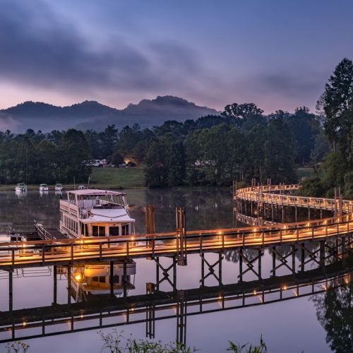 A tranquil lake scene with a lit wooden pier and boat at twilight, surrounded by misty mountains.