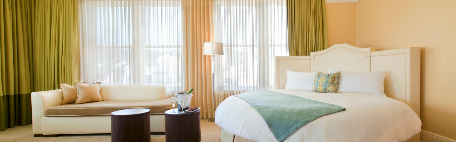 A spacious hotel room with a round bed, green curtains, white sofa, two small round tables, and large windows with sheer drapes.
