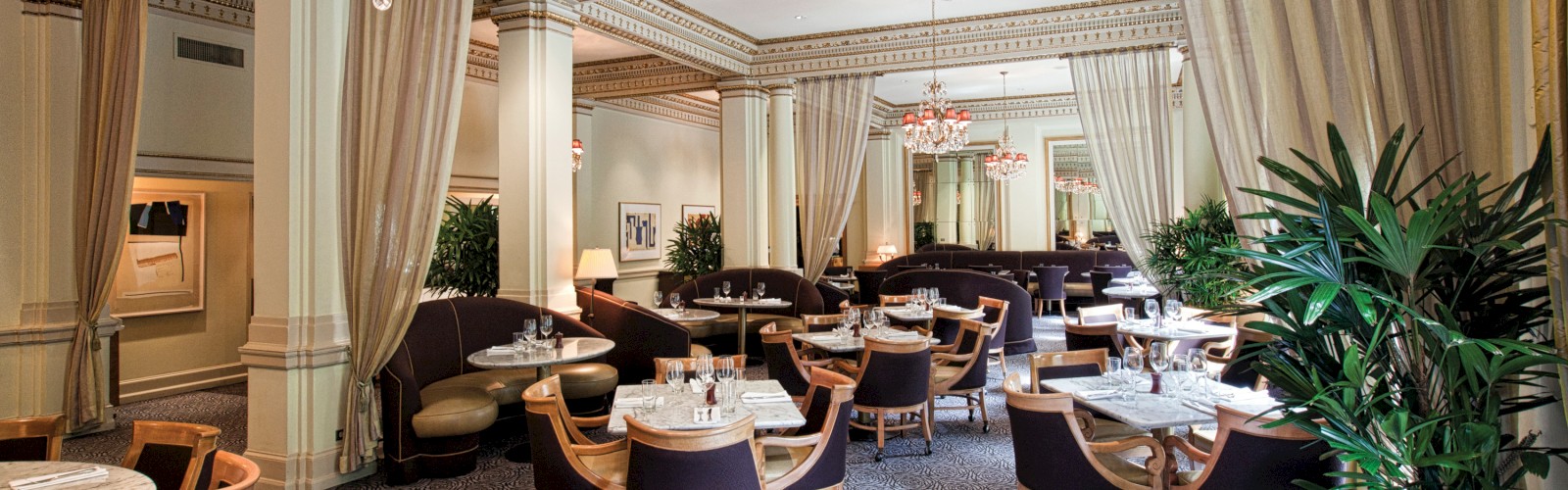 An elegant dining room with chandeliers, round marble tables, and brown chairs arranged neatly, draped curtains, and indoor plants.