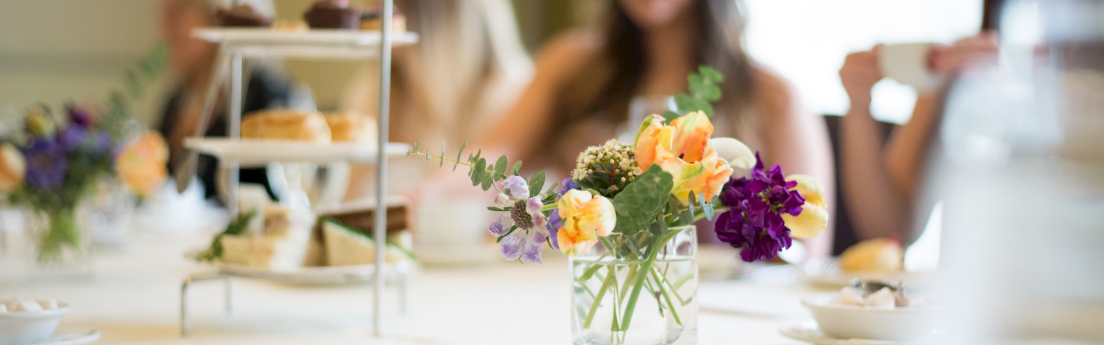 A group of people is sitting at a table set with a vase of flowers, a tiered tray of pastries, and various teacups and plates, enjoying a gathering.