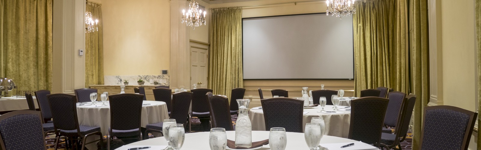The image shows an elegant meeting or conference room with round tables, chairs, a large screen, chandeliers, and papers with pens on the tables.