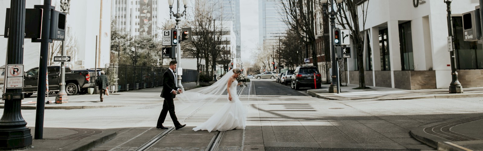 A bride and groom are crossing a city street, holding hands. The scene is urban with tall buildings and streetlights in the background.