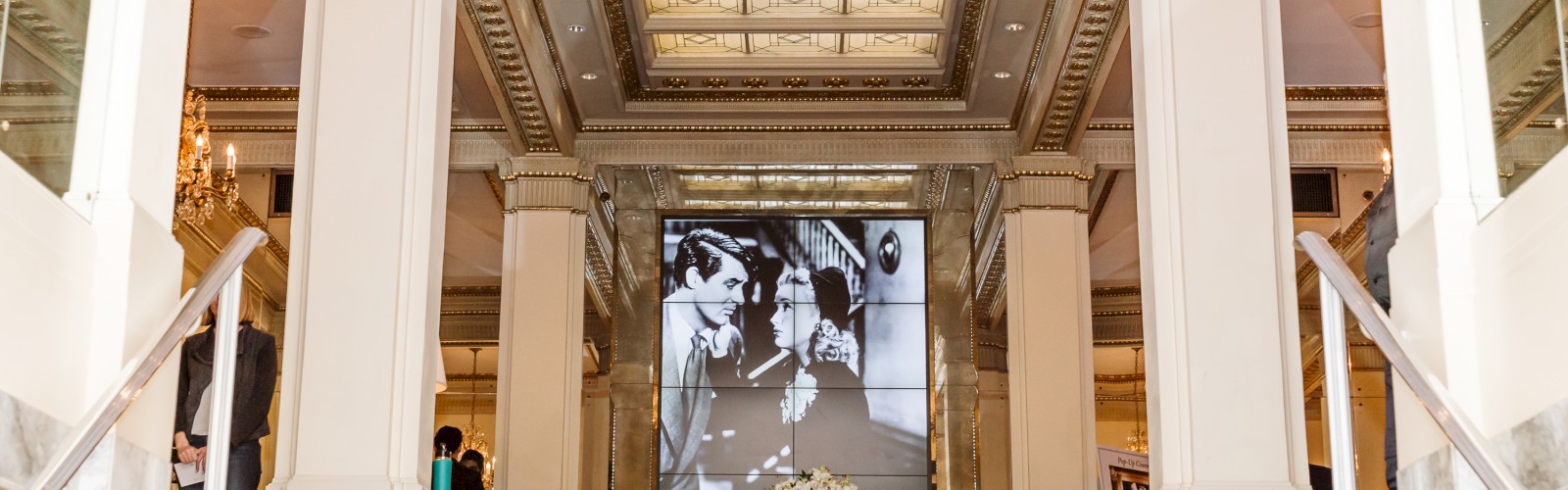 The image shows an ornate interior with tall columns, a chandelier, and a large black-and-white photograph displayed prominently on a screen.