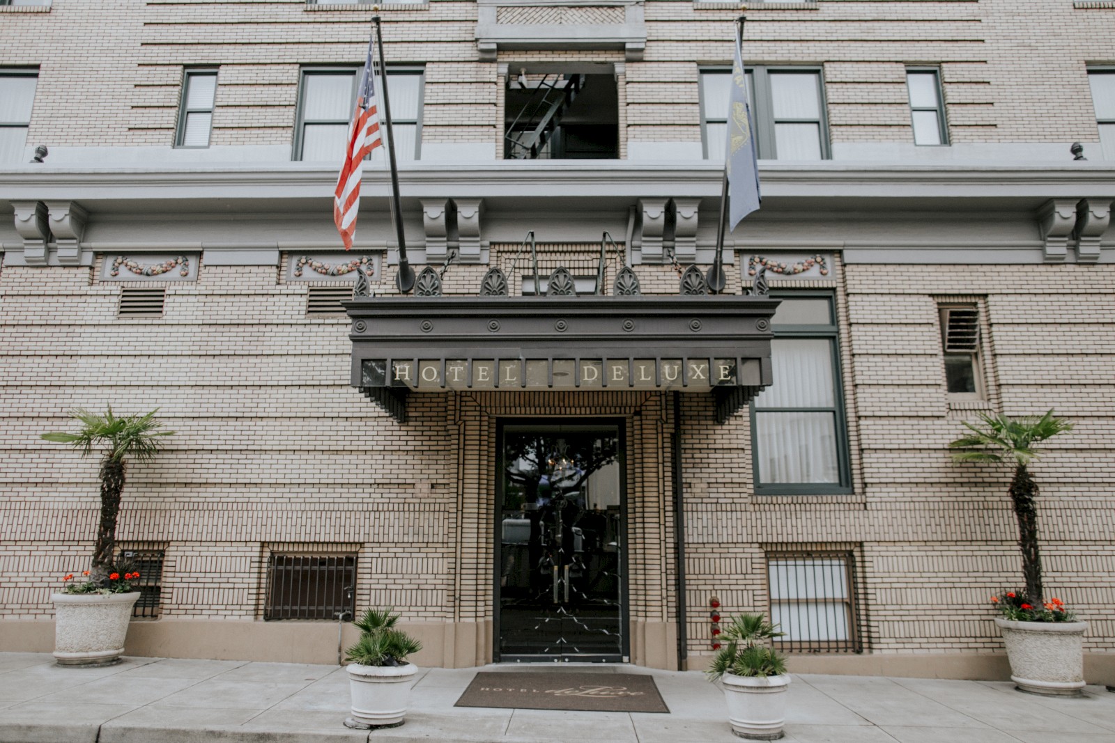The image shows the entrance of Hotel deLuxe, with an American flag and another flag, surrounded by potted plants on both sides.