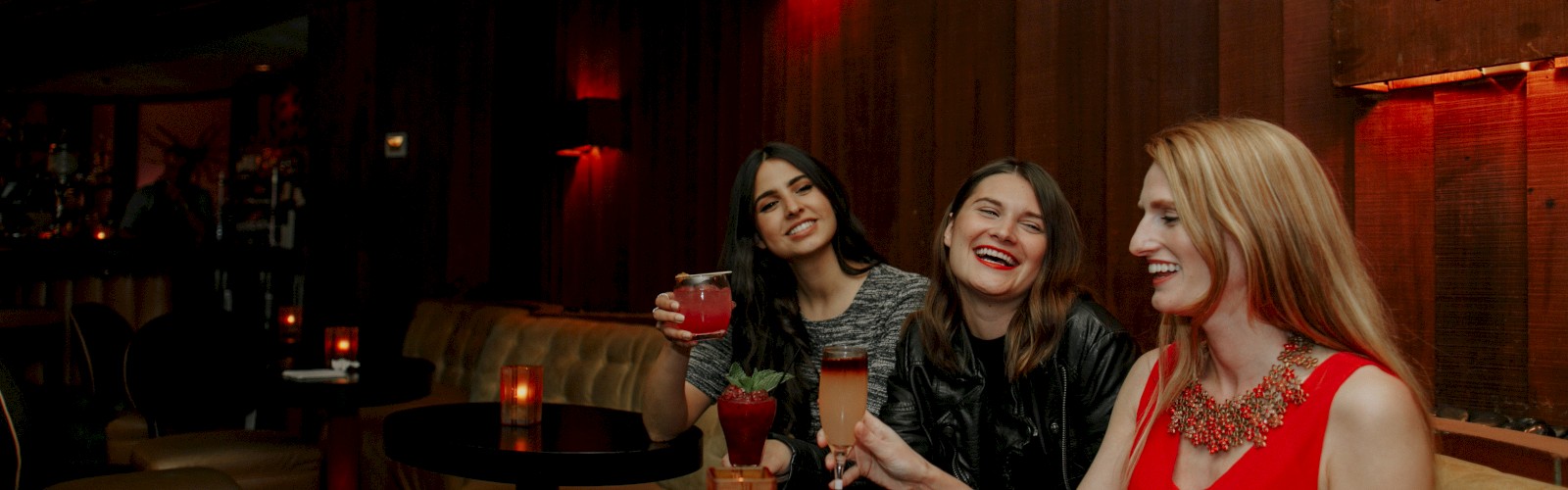 Three women are seated in a dimly lit bar, laughing and holding drinks, with candles on the table.