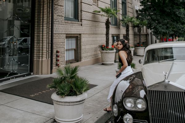 A person in a white dress stands next to a vintage car in front of a stylish building with flags. The entrance has potted plants and a black awning.
