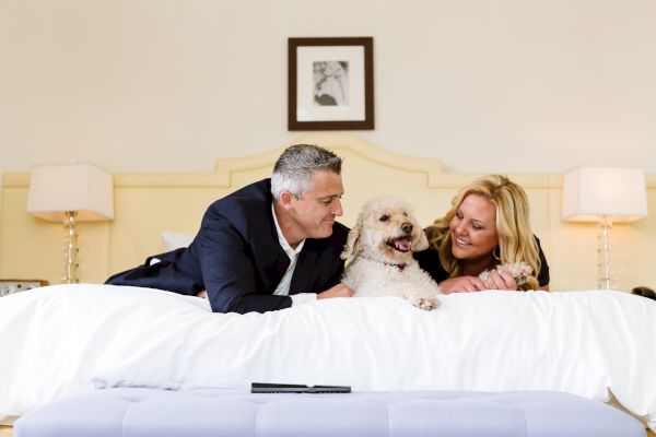 A man and a woman lying on a bed with a fluffy dog between them, smiling at each other.