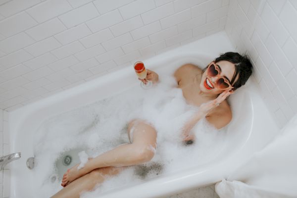 A person is relaxing in a bubble bath, wearing sunglasses, smiling, and holding a glass of presumably champagne.