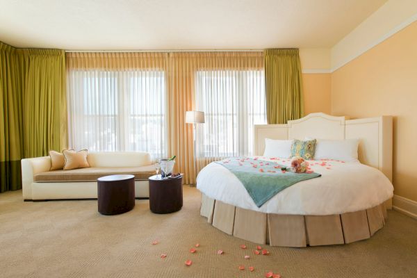 A cozy bedroom with a large bed, a sofa, two round tables, and a window with green curtains. Rose petals are scattered on the floor.