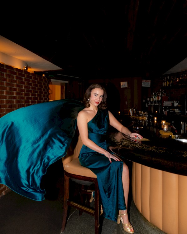 A woman in a flowing blue dress is seated at a bar, holding a drink, with a dramatic pose and a dark, stylish background.