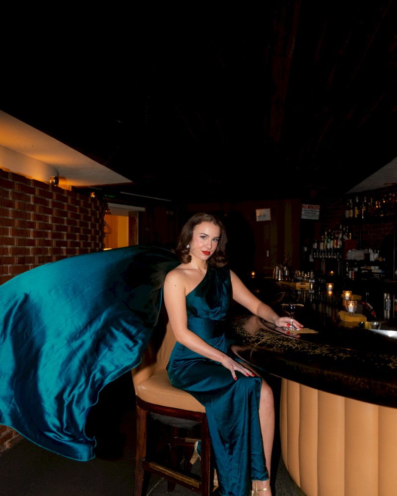 A woman in a flowing blue dress is seated at a bar, holding a drink, with a dramatic pose and a dark, stylish background.