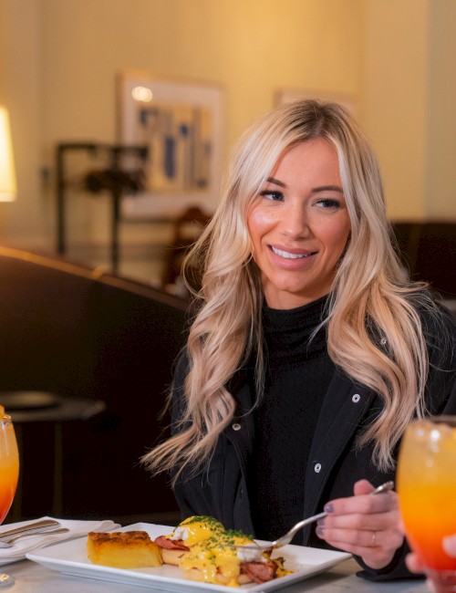 A woman dining at a restaurant, smiling, with plates of food and orange drinks on the table, ends the sentence.