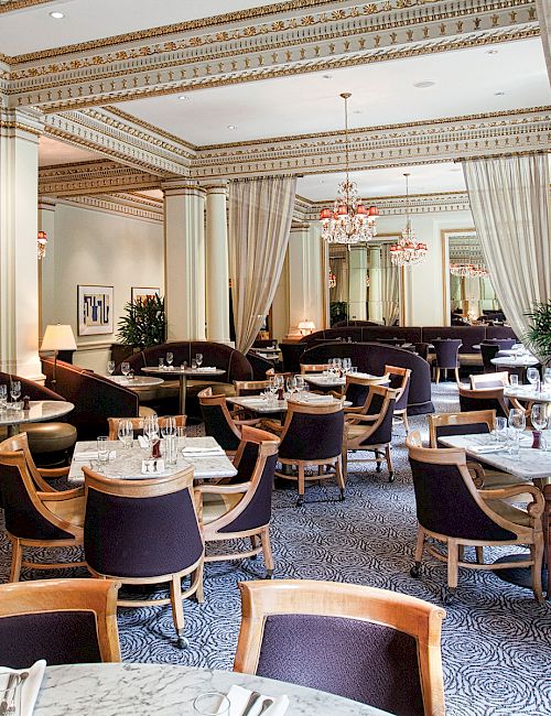 A well-lit, elegant restaurant with marble tables, cushioned chairs, chandeliers, and greenery adorning the interior, creating a luxurious ambiance.