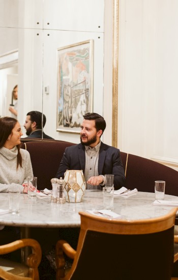 A man and woman sit at a round table in a stylish restaurant, with artwork on the walls and a mirror reflecting the setting.