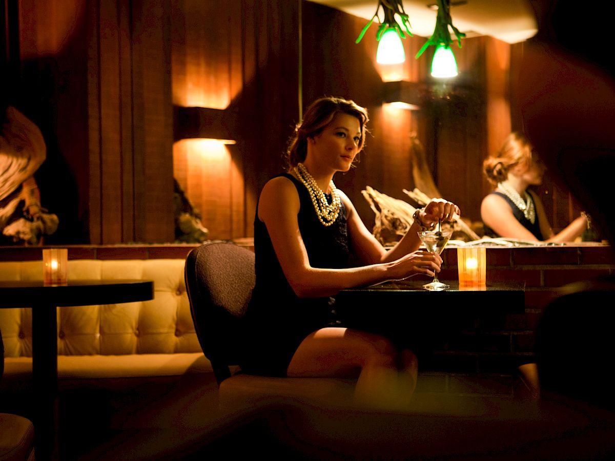 A woman in a dimly lit bar, wearing a black dress and necklace, sits at a table with a drink. She's reflected in a mirror behind her.
