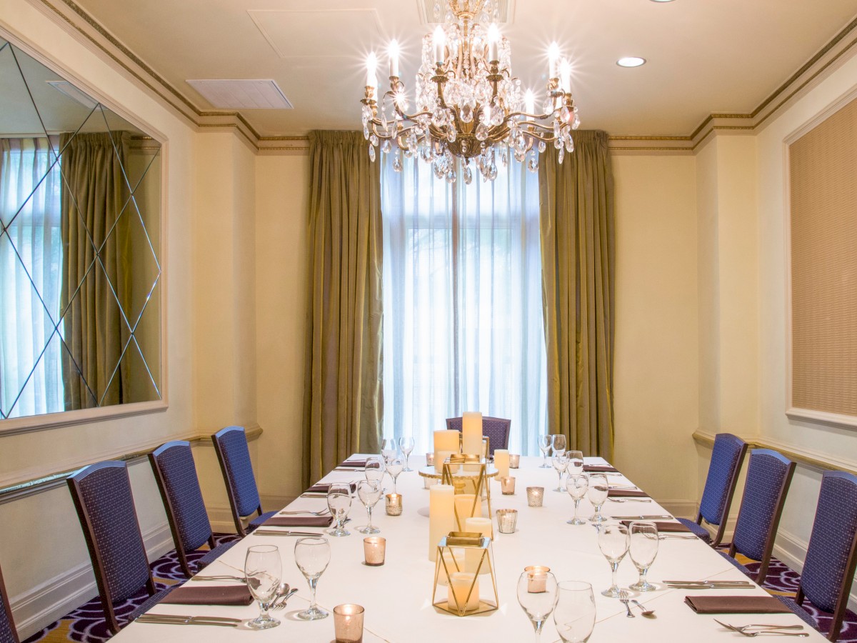 A formal dining room with a long table, set with glassware and napkins. Elegant chandelier and curtains enhance the ambiance.