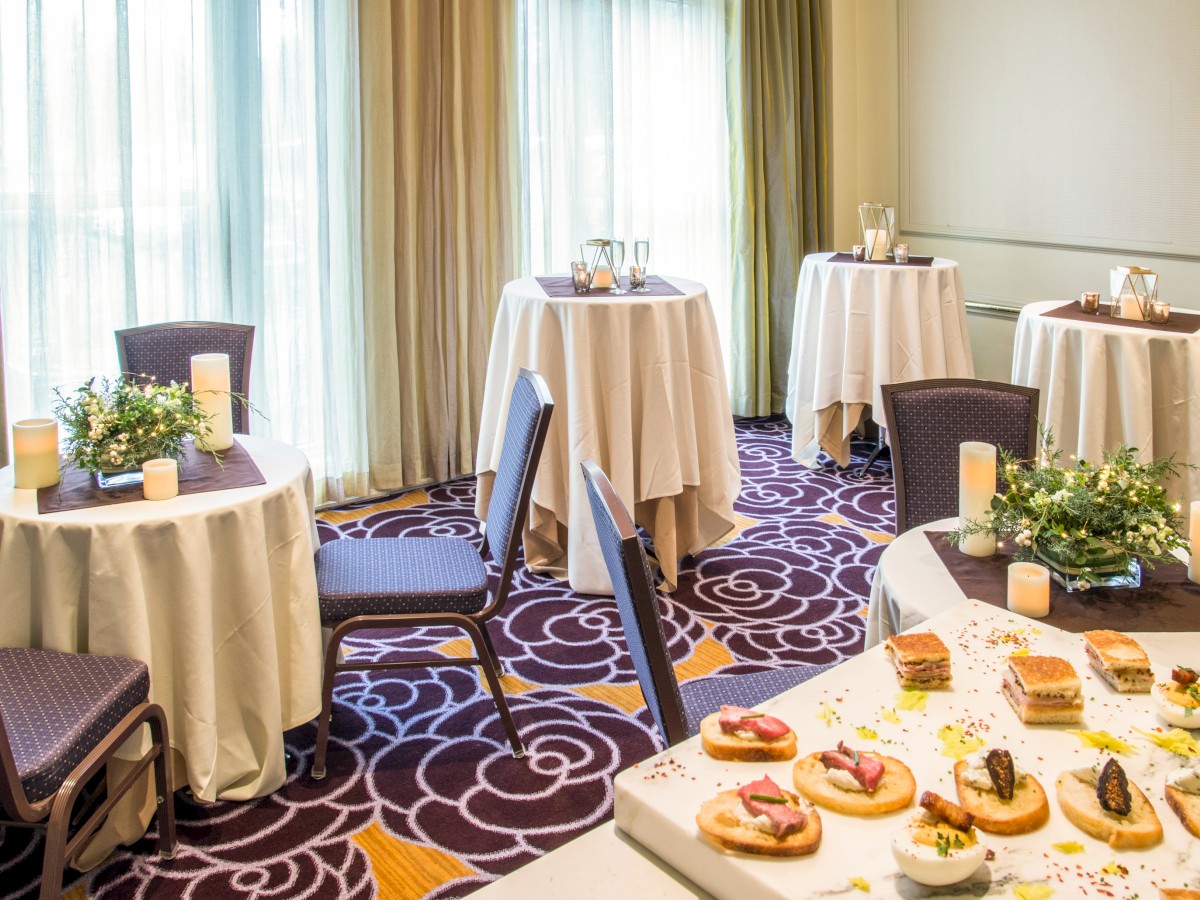 The image shows a decorated room with round tables covered in white cloths, floral centerpieces, candles, chairs, and a small assortment of snacks.