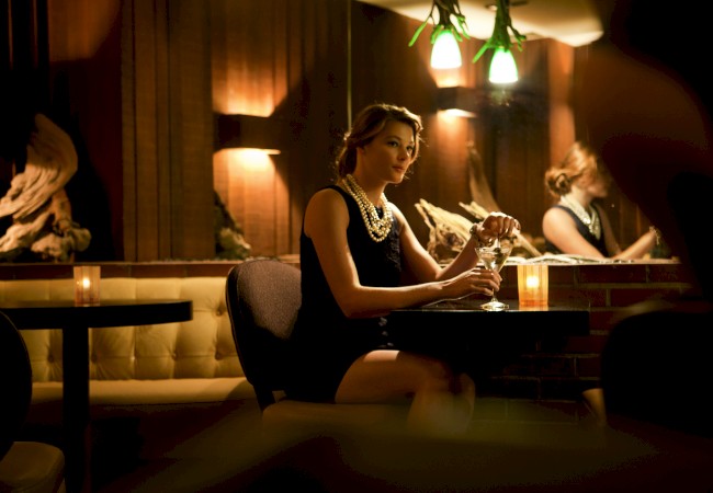 A woman sits alone at a dimly lit bar, holding a glass. She appears thoughtful, and her reflection is visible in the mirror behind her, ending the sentence.