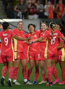 A group of female soccer players in red uniforms huddle together on a field, celebrating with smiles and raised arms.