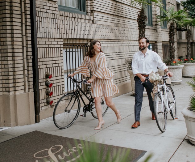 Two people with bicycles stand outside a stylish building with palm trees and a canopy. The sidewalk reads 