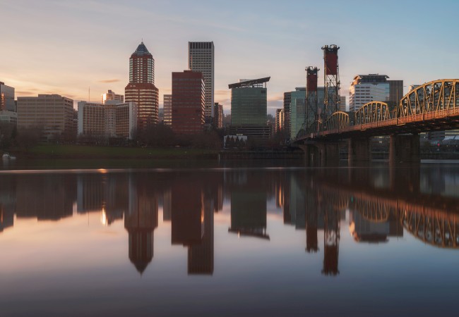 A city skyline reflected in calm water at sunset, with a bridge leading into the city.