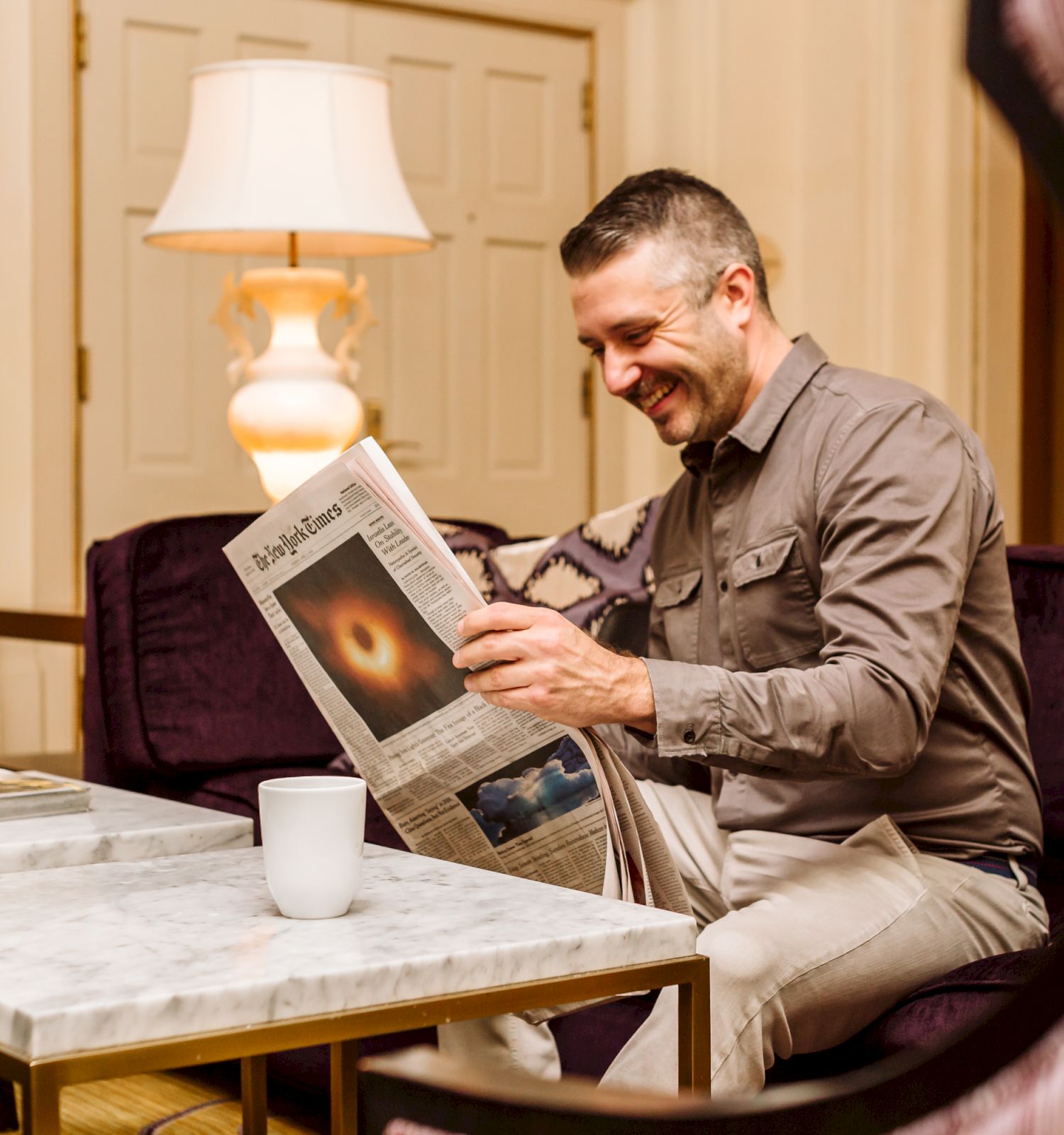 A man sitting in a cozy room is smiling while reading a newspaper at a marble table, with a lamp and coffee cup nearby.
