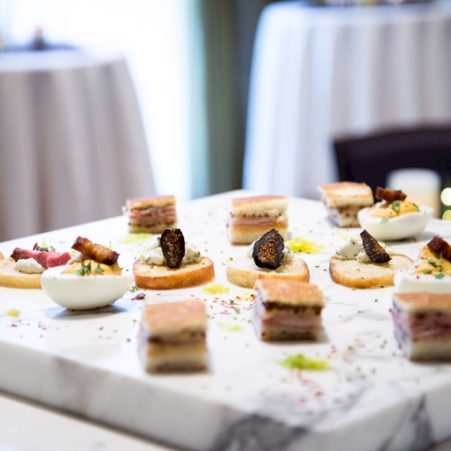 An elegant platter of assorted hors d'oeuvres, including sandwiches and small bites, on a marble surface with a glass of sparkling drink nearby.