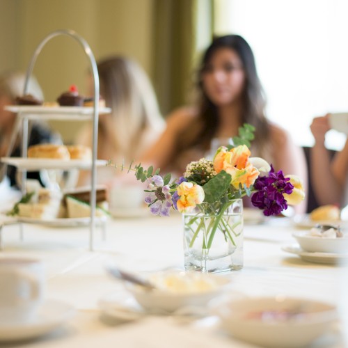 The image shows a table set for tea with flowers, cups, and a tiered tray of pastries, while blurred people sit in the background conversing.