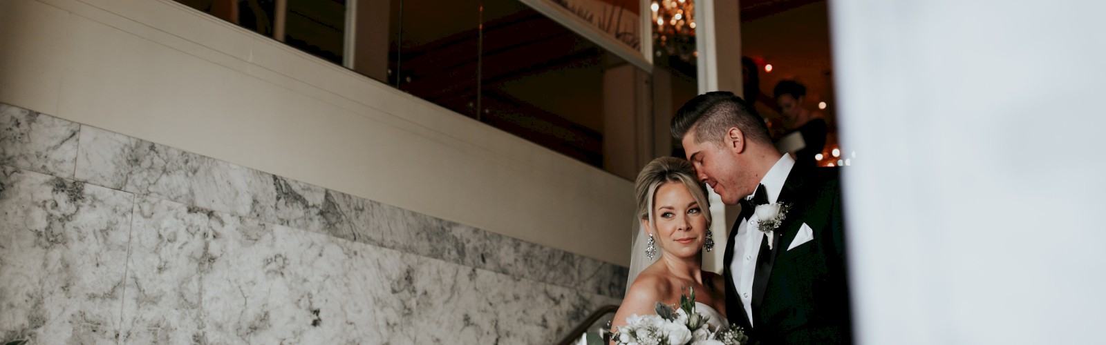 A bride and groom embrace on a staircase, the bride holding a bouquet of flowers, with decorative artwork and chandeliers in the background.