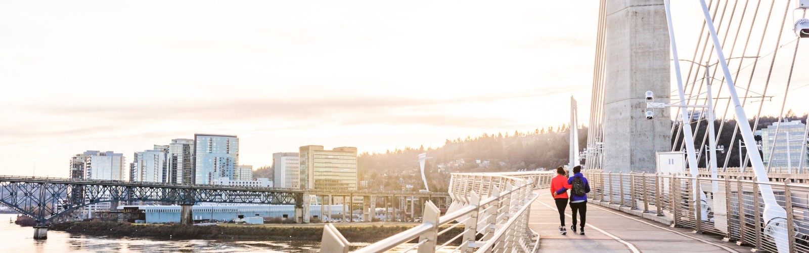 Two people walk on a modern bridge with cables, overlooking a river and a cityscape with buildings and a distant hill at sunset.