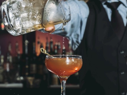 A bartender pours a cocktail from a mixing glass into a coupe glass with an olive garnish.