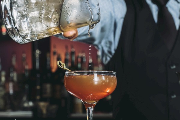 A bartender pours a cocktail from a mixing glass into a coupe glass with an olive garnish.