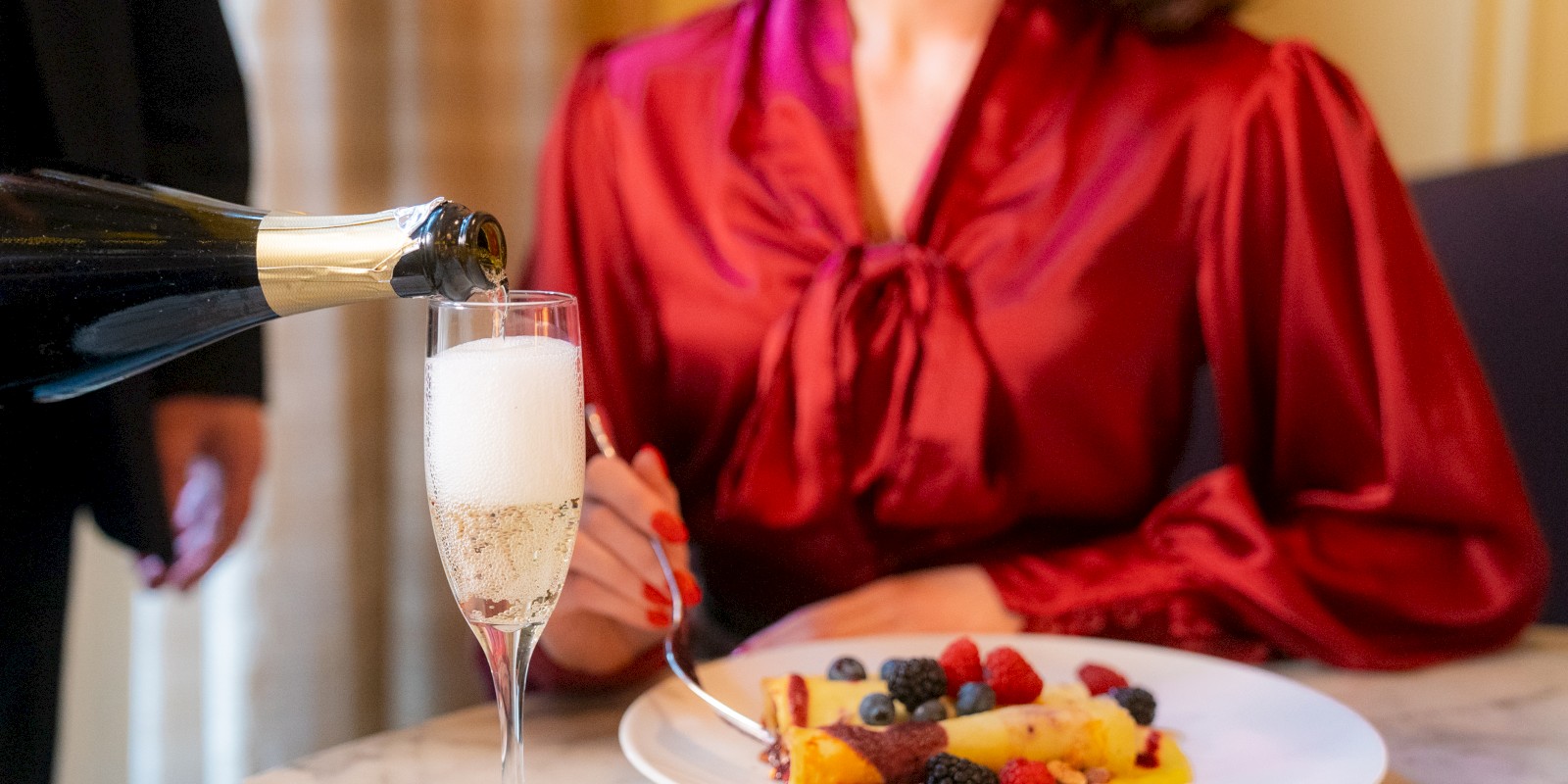 A person in a red blouse is sitting at a table with a dessert and being served champagne in a glass.