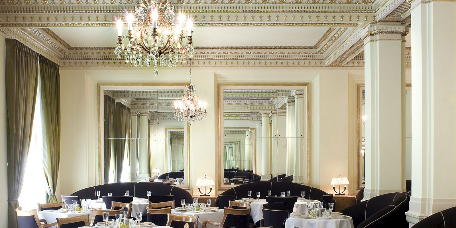 Elegant dining room with tables set, chandelier above, ornate ceiling details.