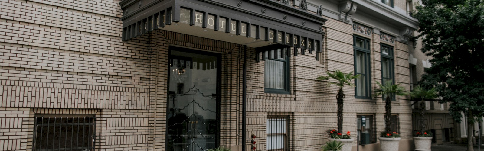 The image shows the entrance to a brick building with a decorative awning, flags, and potted plants along the sidewalk.