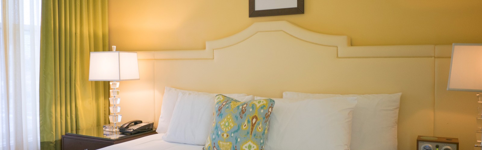 A well-made bed in a cozy room with yellow walls, green curtains, bedside lamps, and a decorative headboard, adorned with a patterned cushion.