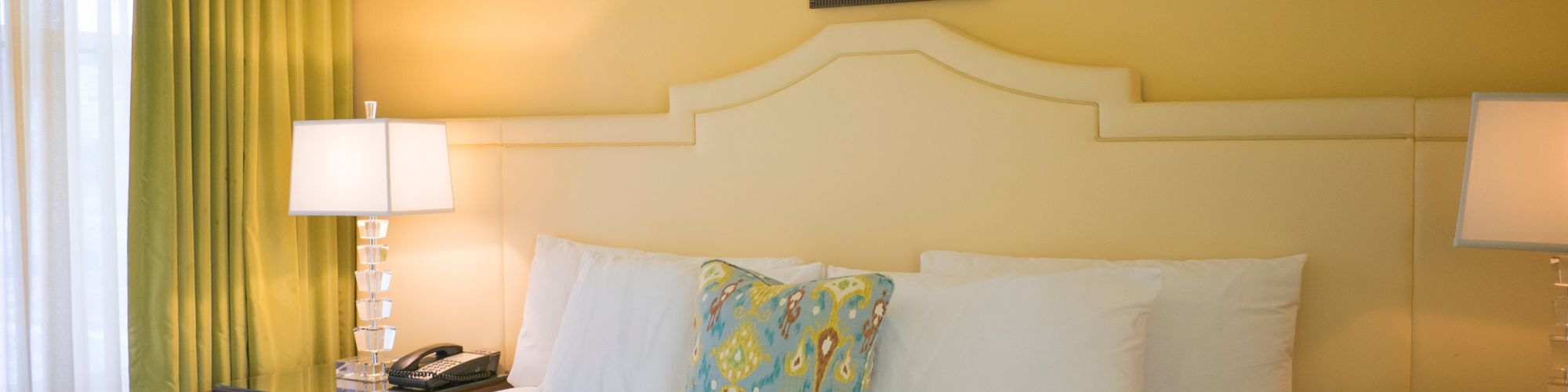 A well-made bed in a cozy room with yellow walls, green curtains, bedside lamps, and a decorative headboard, adorned with a patterned cushion.