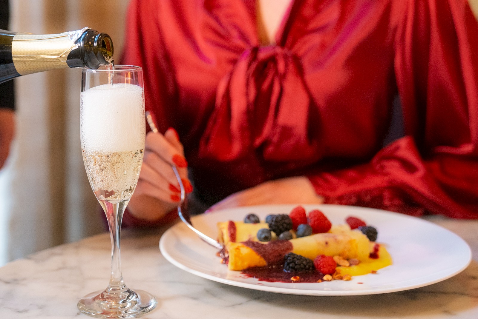 A person in a red blouse with a dessert plate is served champagne in a glass, featuring various berries on a table.