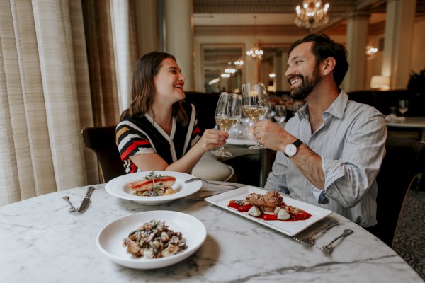 A couple sits at a restaurant table, toasting with wine glasses and smiling, with plates of food in front of them.