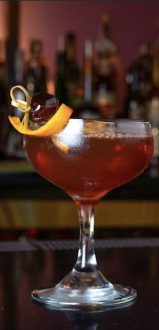 A cocktail in a coupe glass, garnished with an orange twist, on a dimly lit bar counter with bottles in the background.