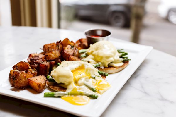 A plate with eggs benedict, asparagus, and potato cubes served with a side of sauce, on a marble countertop by a window.