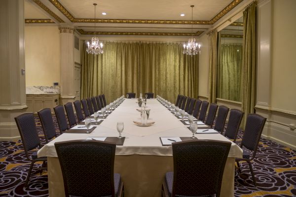 A conference room with a long table arranged in a U-shape, set with glasses, notepads, and pens, featuring chandeliers and elegant decor.