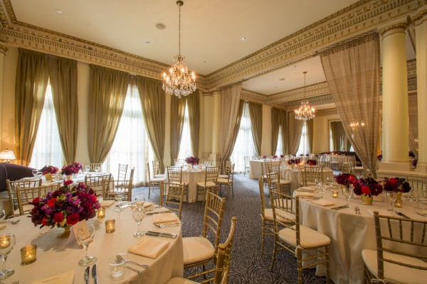A luxurious banquet hall with elegant chandeliers, round tables with white tablecloths, flower centerpieces, and gold chairs set for an event.